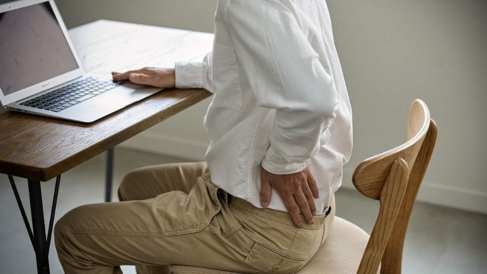 Office worker sitting at a desk with a laptop while holding their lower back in pain, showing how prolonged sitting causes lower back pain that a chiropractor can treat
