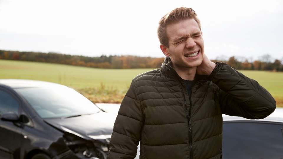 Man holding his neck in pain while standing near a damaged car, representing common injuries after a car accident