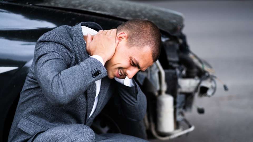 Man grimacing and holding his neck in pain beside a damaged car after a collision, showing why seeing an auto accident chiropractor near me is critical for whiplash treatment