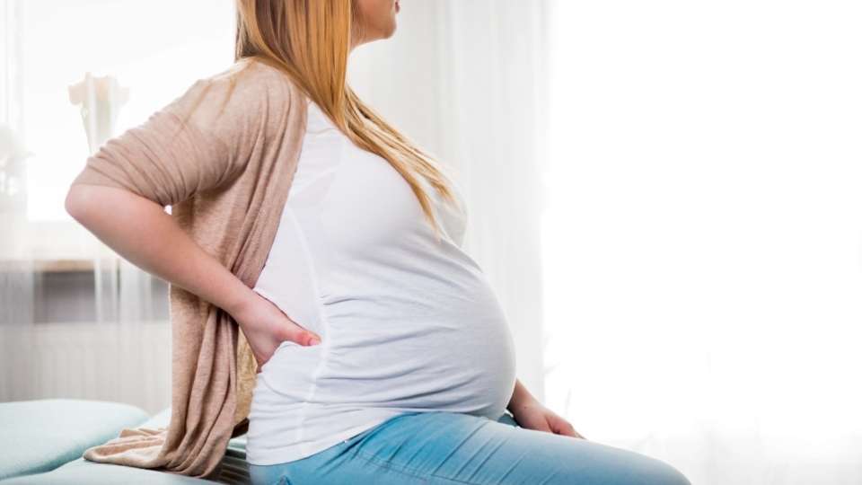 Pregnant woman sitting on a bed and holding her lower back in pain, illustrating prenatal back pain during pregnancy