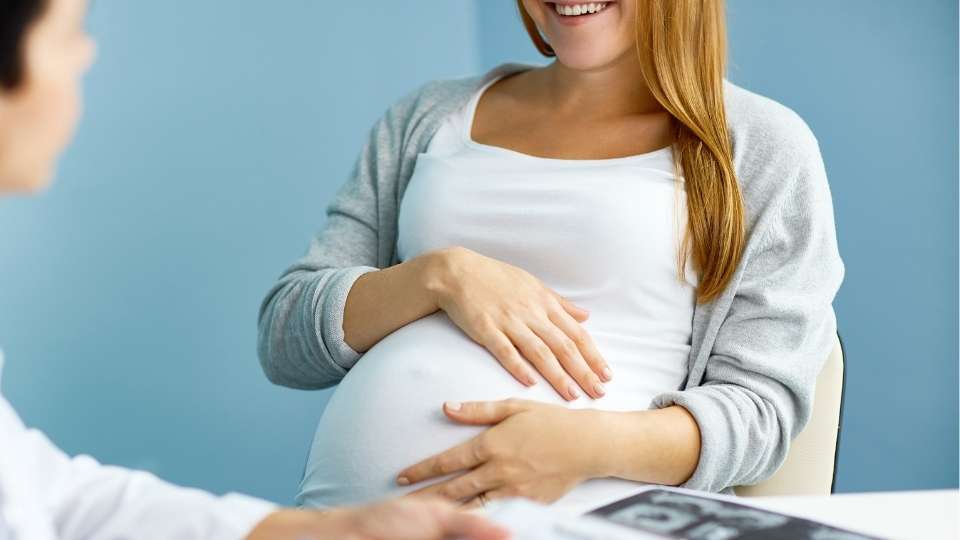 Smiling pregnant woman cradling her belly during a prenatal chiropractic consultation with her doctor
