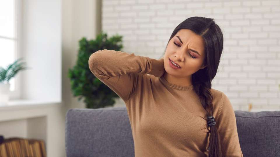 A woman seated on a couch clutching the back of her neck in pain, representing chronic neck pain from long-term untreated whiplash