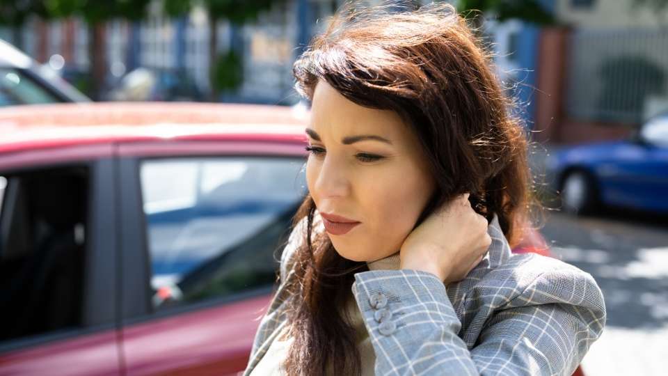 A woman standing near a car on a street and holding the back of her neck in pain, illustrating whiplash symptoms following a rear-end collision 