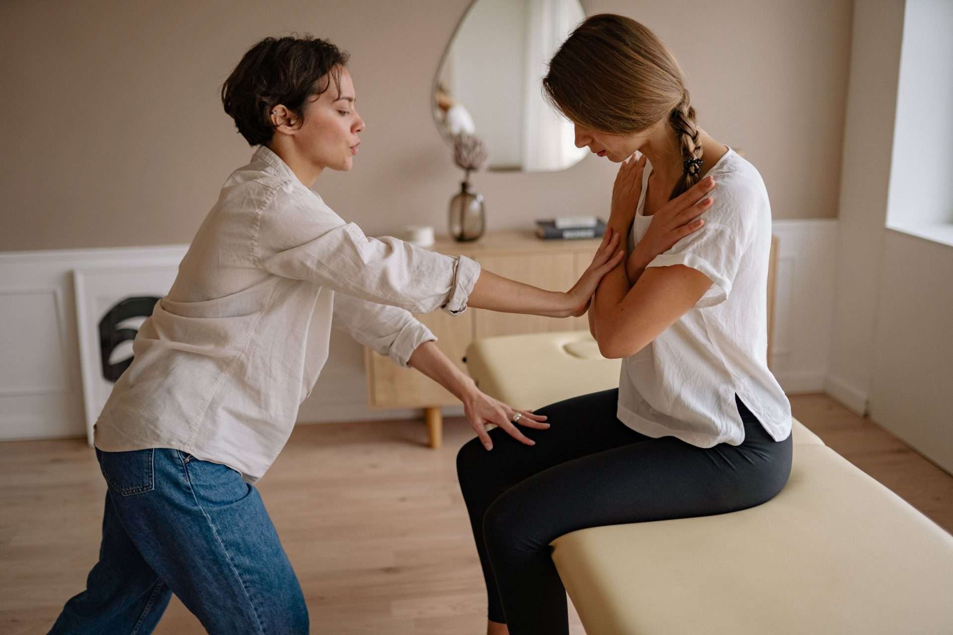 A practitioner guides a seated woman with crossed arms through a hands-on upper back technique on a padded treatment table.