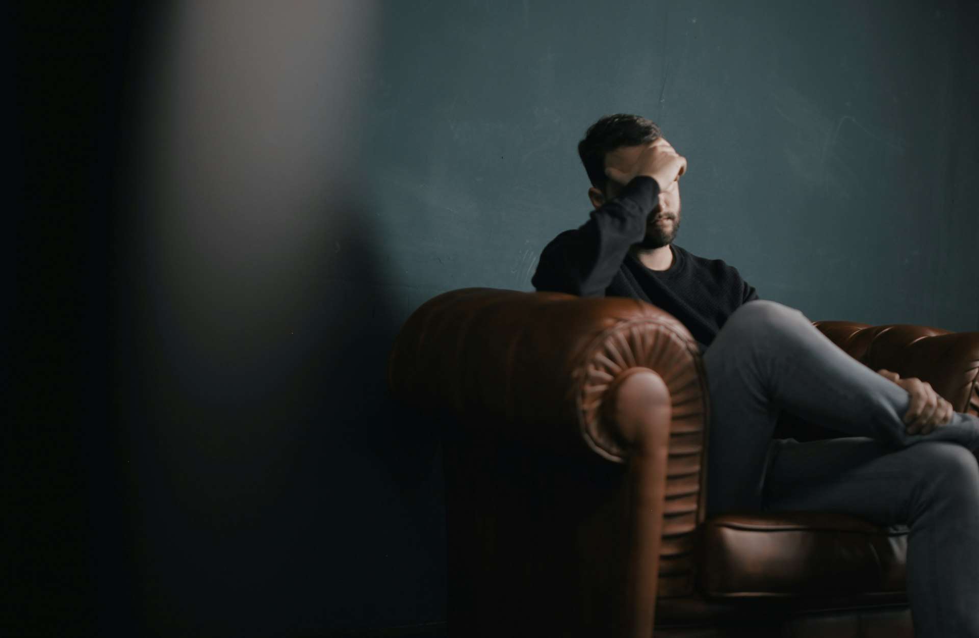 A man sits on a leather sofa with one hand on his head, a quiet moment many people know during a slow concussion recovery.