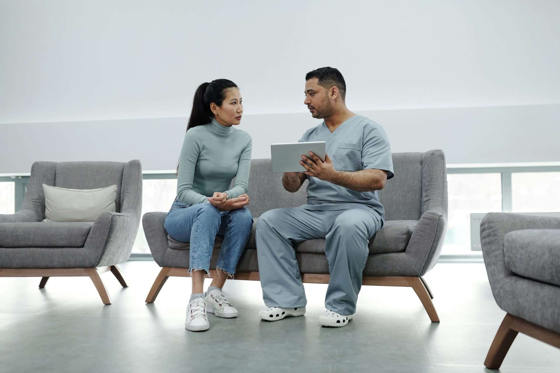 A clinician in scrubs holds a tablet and talks with a seated woman, reviewing the health history behind lingering symptoms.
