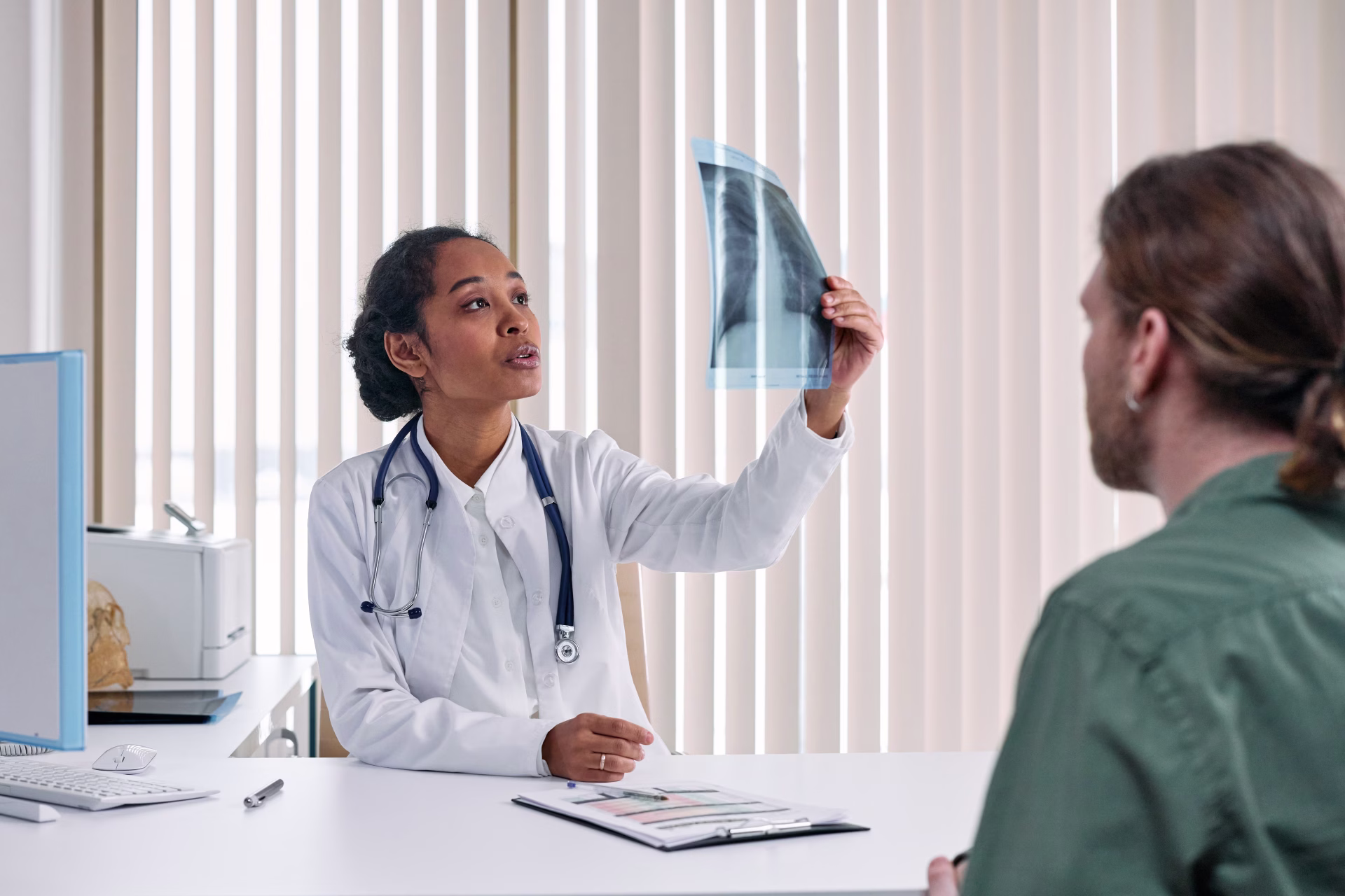 An injury doctor in a white coat holds up an X-ray while talking with a seated patient about what the film shows.