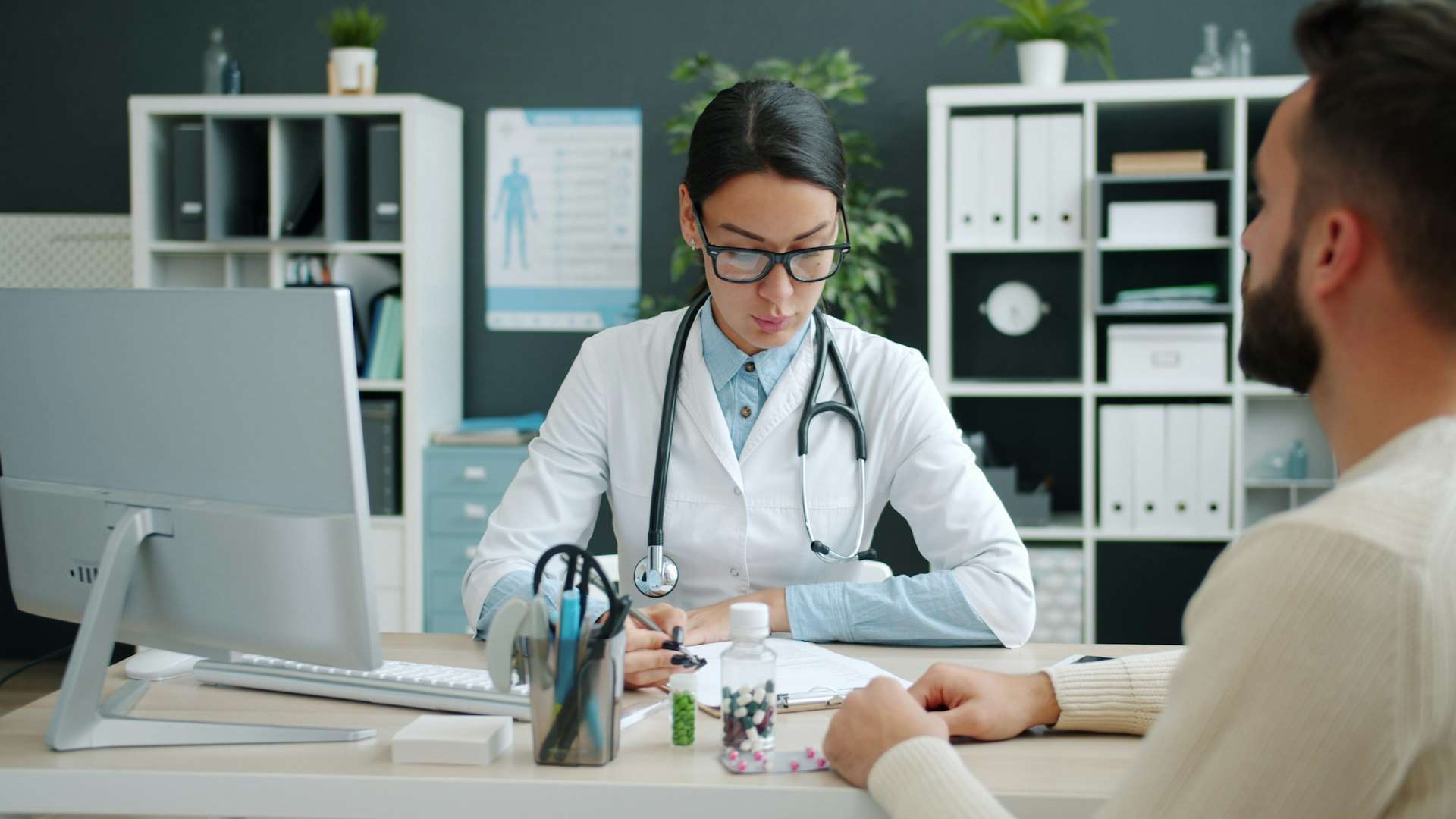 A clinician writes notes at her desk while a patient describes how his injury happened during a first consultation visit.
