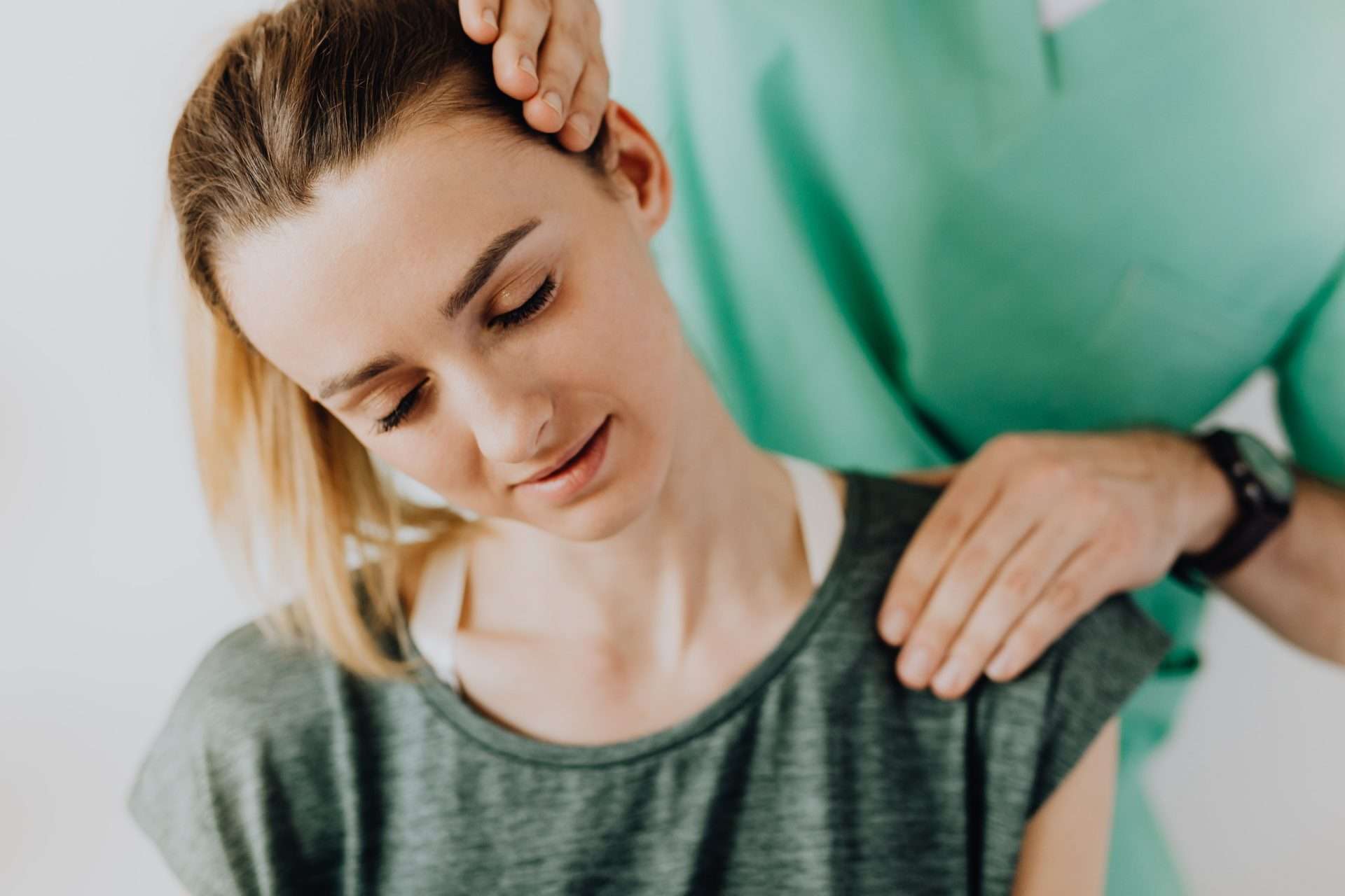 A clinician tilts a seated woman's head to one side to stretch her neck and shoulder during a neck pain treatment visit.