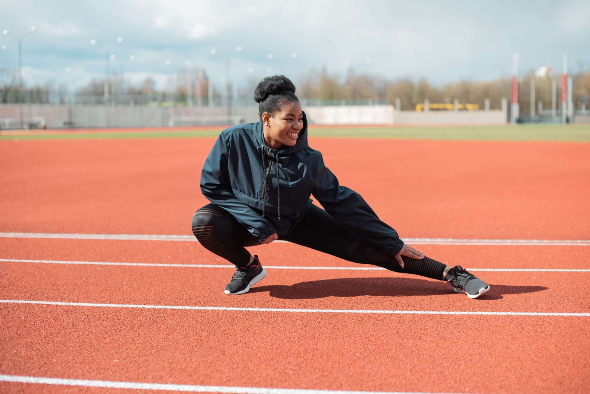 A smiling woman in a black jacket and tights holds a deep side lunge on a red running track before a training run.