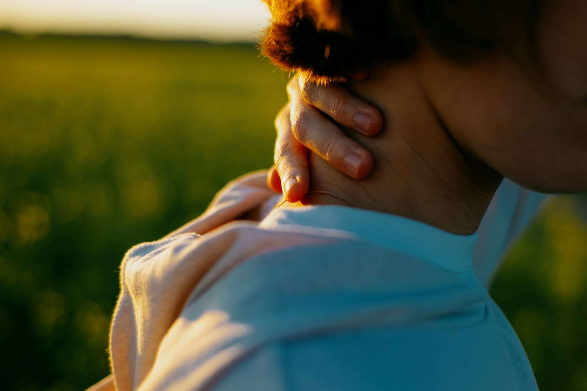 Woman in a light shirt holds the back of her sore neck in a sunlit field, unsure whether to see a spine specialist yet.