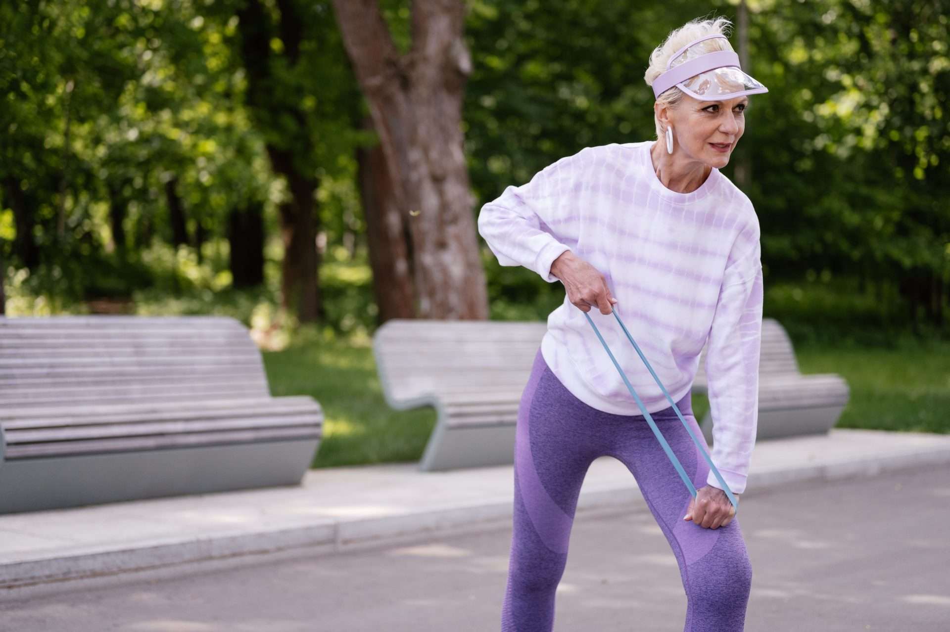 An older woman in a lavender sweater pulls a resistance band in a park, building strength around her knees and hips.