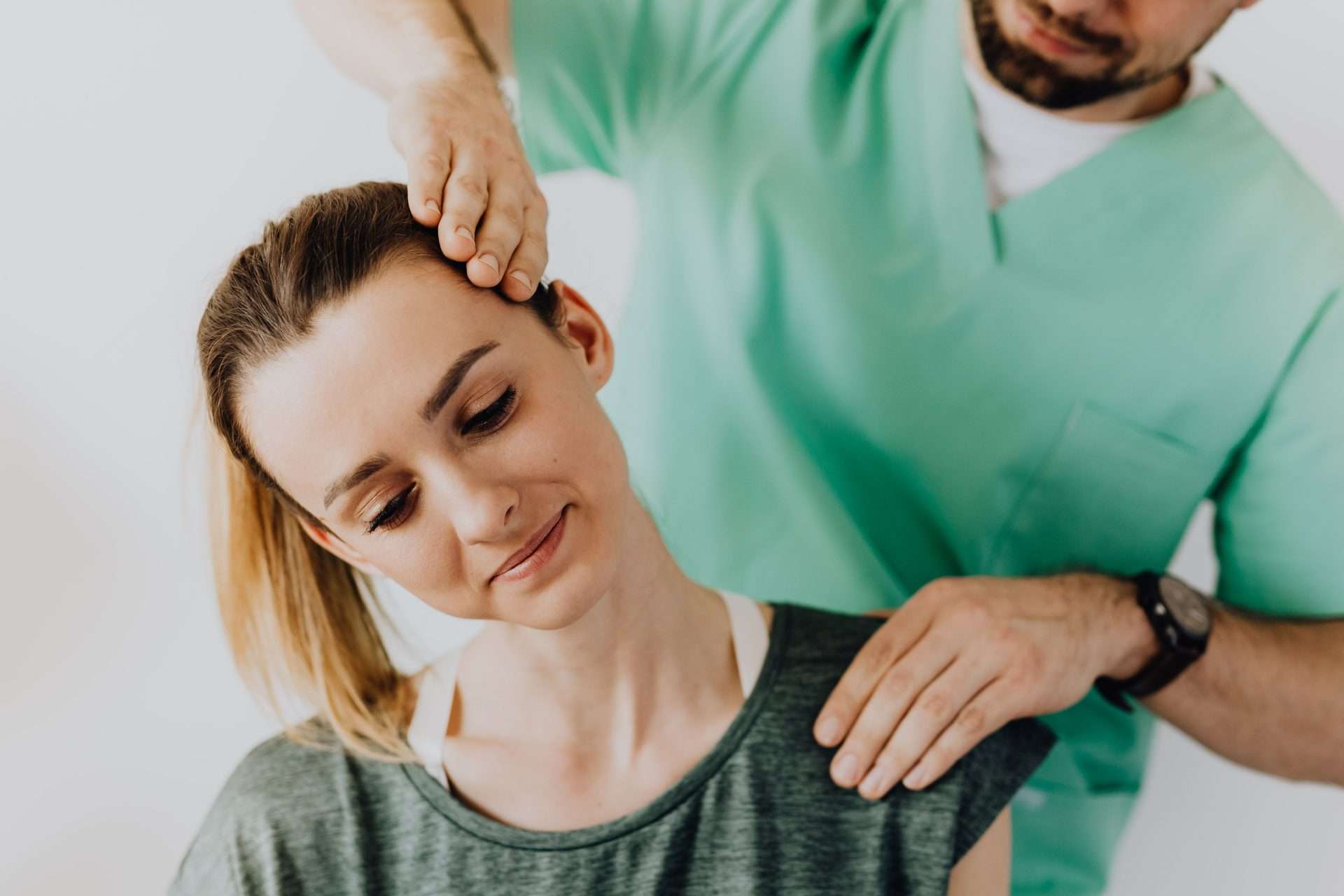 Clinician in green scrubs tilts a woman's head to one side to test neck motion during an exam for neck strain after a crash.